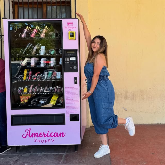 Máquina rosa de American Shopps con perfumes y cremas, frente a una pared amarilla.